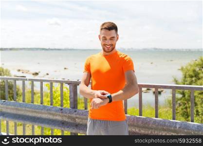 fitness, sport, people, technology and healthy lifestyle concept - smiling young man with smart wristwatch at summer seaside