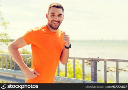 fitness, sport, people, technology and healthy lifestyle concept - smiling young man with heart rate watch running at summer seaside