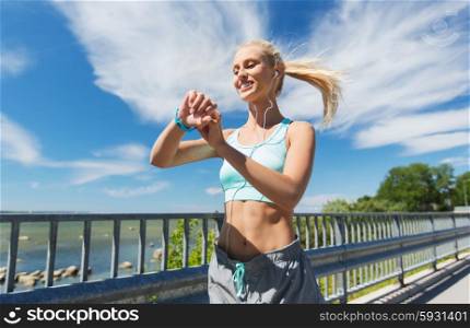 fitness, sport, people, technology and healthy lifestyle concept - smiling young woman with heart rate watch and earphones exercising outdoors