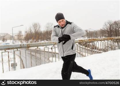 fitness, sport, people, technology and healthy lifestyle concept - young man in earphones running along snow covered winter bridge and looking at activity tracker or smart watch