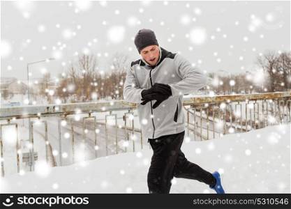 fitness, sport, people, technology and healthy lifestyle concept - young man in earphones running along snow covered winter bridge and looking at activity tracker or smart watch. man with earphones running along winter bridge