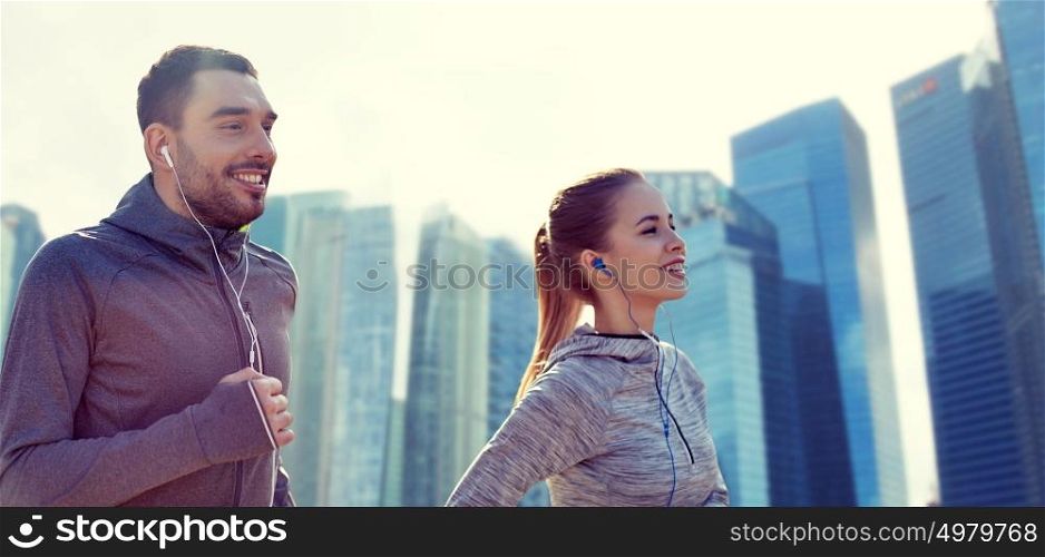 fitness, sport, people, technology and lifestyle concept - happy couple running and listening to music in earphones over singapore city skyscrapers background. happy couple with earphones running in city
