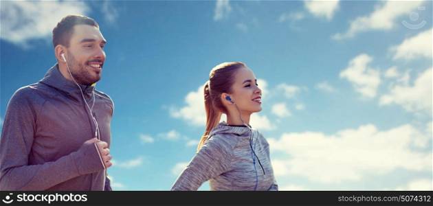 fitness, sport, people, technology and lifestyle concept - happy couple running and listening to music in earphones over blue sky and clouds background. happy couple with earphones running over blue sky