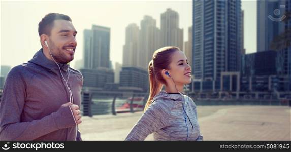 fitness, sport, people, technology and lifestyle concept - happy couple running and listening to music in earphones over dubai city street background. happy couple with earphones running in city