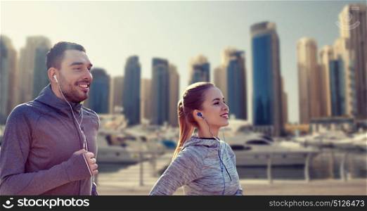 fitness, sport, people, technology and lifestyle concept - happy couple running and listening to music in earphones over dubai city street background. happy couple with earphones running in city