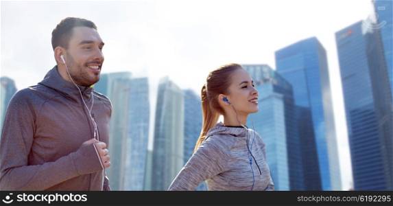 fitness, sport, people, technology and lifestyle concept - happy couple running and listening to music in earphones over singapore city skyscrapers background