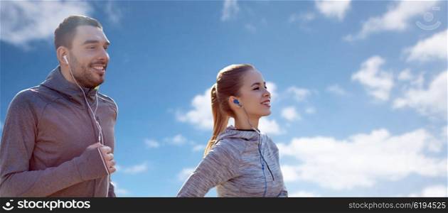 fitness, sport, people, technology and lifestyle concept - happy couple running and listening to music in earphones over blue sky and clouds background
