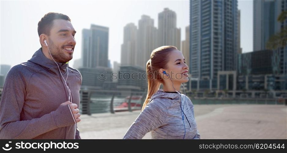 fitness, sport, people, technology and lifestyle concept - happy couple running and listening to music in earphones over dubai city street background