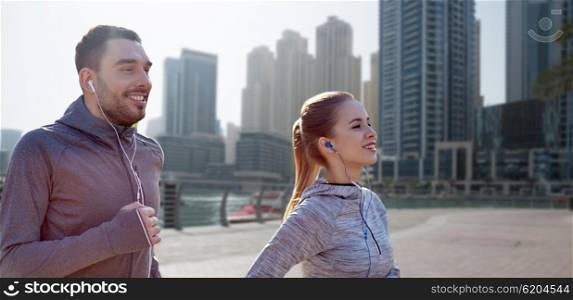 fitness, sport, people, technology and lifestyle concept - happy couple running and listening to music in earphones over dubai city street background