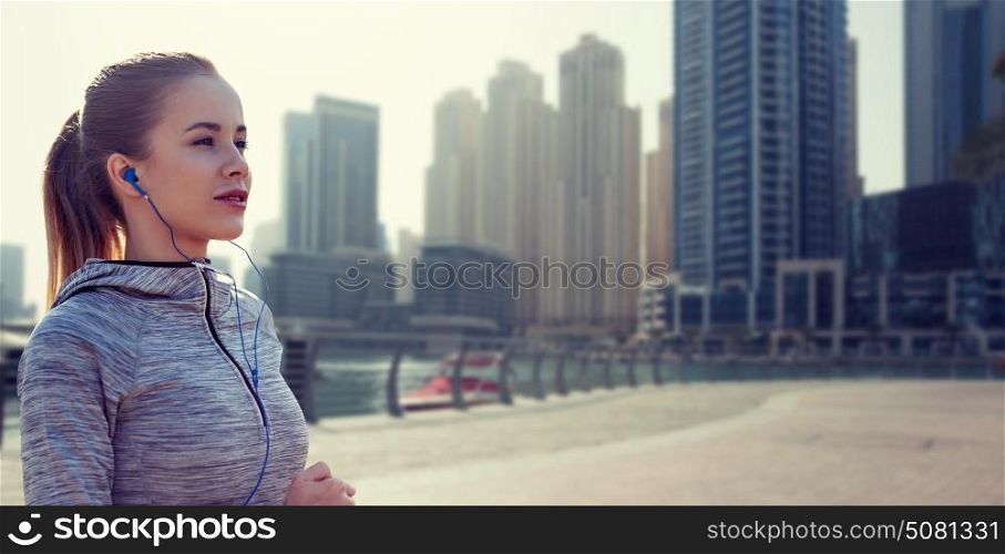 fitness, sport, people, technology and lifestyle concept - happy woman running and listening to music in earphones over dubai city street background. happy woman with earphones running over dubai city