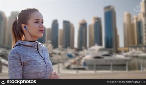 fitness, sport, people, technology and lifestyle concept - happy woman running and listening to music in earphones over dubai city street background