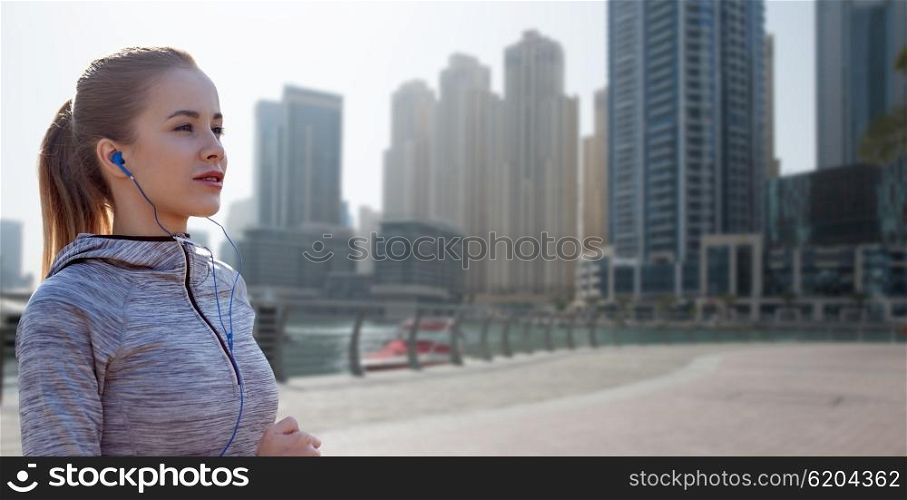 fitness, sport, people, technology and lifestyle concept - happy woman running and listening to music in earphones over dubai city street background