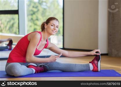 fitness, sport, people, technology and lifestyle concept - smiling woman with smartphone and earphones stretching in gym
