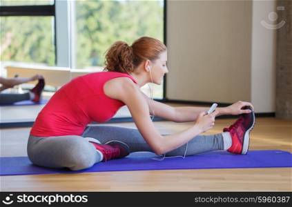 fitness, sport, people, technology and lifestyle concept - smiling woman with smartphone and earphones stretching in gym