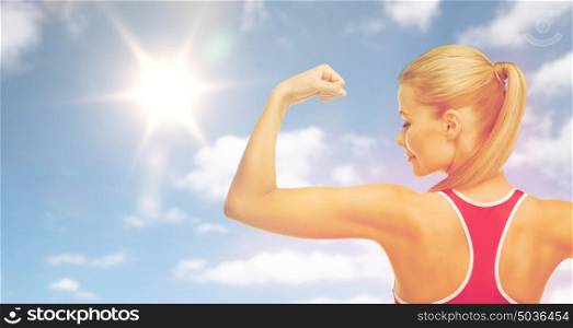 fitness, sport, power, strength and people concept - happy sporty young woman showing biceps over sky and sun background. happy sporty woman showing biceps over sky and sun