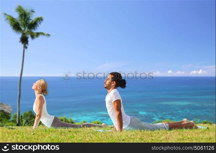 fitness, sport, relax and people concept - couple making yoga cobra pose over natural background over exotic natural background with palm tree and ocean. couple making yoga cobra pose outdoors