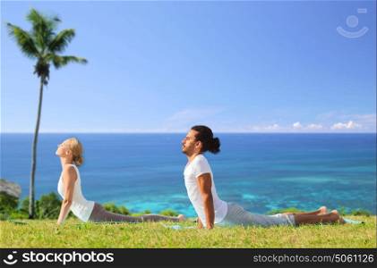 fitness, sport, relax and people concept - couple making yoga cobra pose over natural background over exotic natural background with palm tree and ocean. couple making yoga cobra pose outdoors