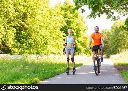 fitness, sport, summer, people and healthy lifestyle concept - happy couple with rollerblades and bicycle riding outdoors at summer