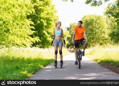 fitness, sport, summer, people and healthy lifestyle concept - happy couple with roller skates and bicycle riding and talking outdoors at summer