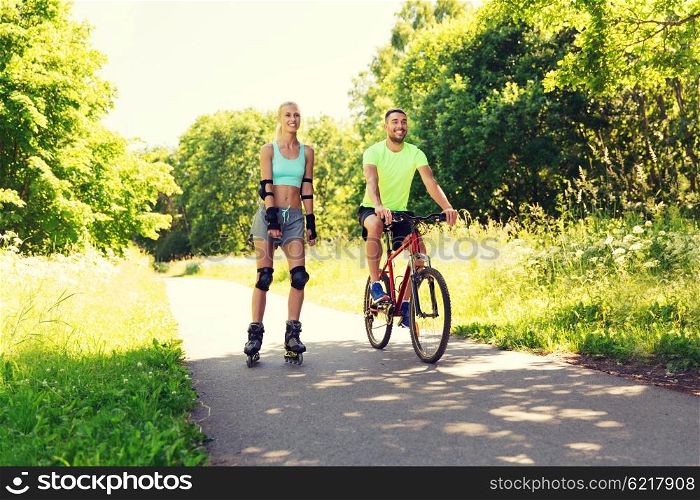 fitness, sport, summer, people and healthy lifestyle concept - happy couple with rollerblades and bicycle riding outdoors at summer