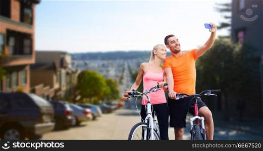 fitness, sport, technology and healthy lifestyle concept - happy couple riding bicycles and taking selfie by smartphone over san francisco city background. couple with bicycles taking selfie by smartphone. couple with bicycles taking selfie by smartphone