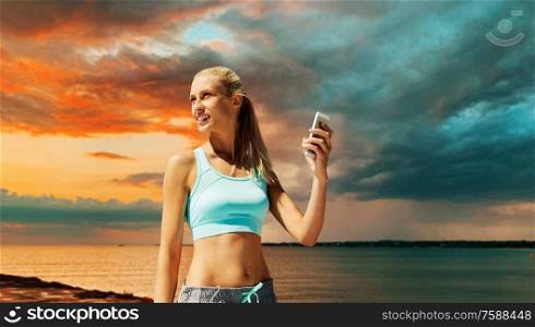 fitness, sport, technology and people concept - smiling young woman with smartphone exercising over sea and sunset sky on background. happy woman with smartphone exercising over sea