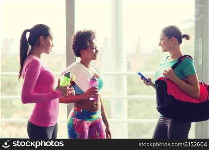 fitness, sport, training and lifestyle concept - group of happy women with bottles of water, smartphone and bag talking in gym