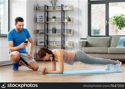 fitness, sport, training and lifestyle concept - happy smiling personal trainer with smartphone and woman doing plank at home. woman with personal trainer doing plank at home