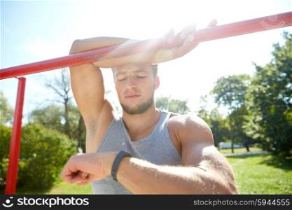 fitness, sport, training and lifestyle concept - happy young man looking at heart-rate watch bracelet and exercising on horizontal bar in summer park