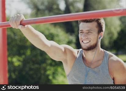 fitness, sport, training and lifestyle concept - happy young man with earphones listening to music and exercising on horizontal bar outdoors