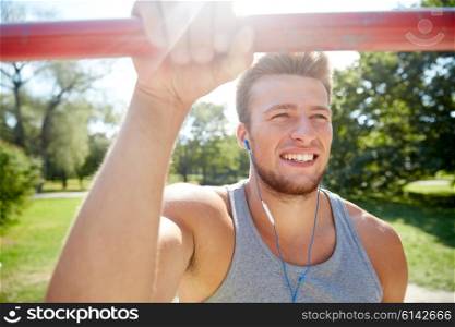 fitness, sport, training and lifestyle concept - happy young man with earphones listening to music and exercising on horizontal bar outdoors