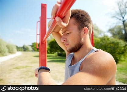 fitness, sport, training and lifestyle concept - young man looking at heart-rate watch bracelet and exercising on horizontal bar outdoors