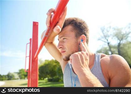 fitness, sport, training and lifestyle concept - young man with earphones listening to music and exercising on horizontal bar outdoors