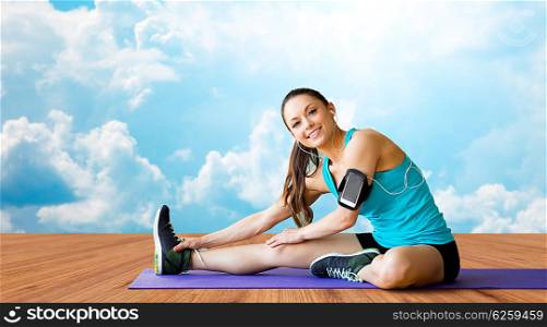 fitness, sport, training and people concept - smiling woman with smartphone and earphones listening to music and stretching leg on exercise mat over wooden floor and sky with white clouds background