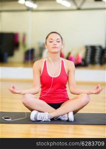 fitness, sport, training, gym, technology and lifestyle concept - smiling teenage girl with smartphone and earphones meditating in gym