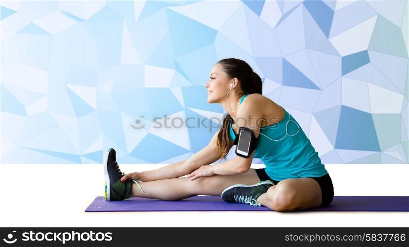 fitness, sport, training, technology and people concept - smiling woman with smartphone and earphones listening to music and stretching leg on exercise mat over blue low poly background