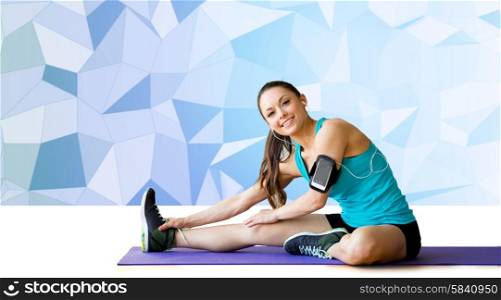 fitness, sport, training, technology and people concept - smiling woman with smartphone and earphones listening to music and stretching leg on exercise mat over blue low poly background