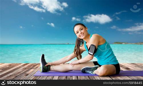 fitness, sport, training, technology and people concept - smiling woman with smartphone and earphones listening to music and stretching leg over sea and wooden berth at resort background