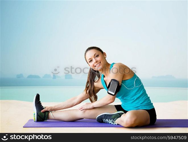 fitness, sport, training, technology and people concept - smiling woman with smartphone and earphones listening to music and stretching leg on exercise mat over sea and pool at hotel resort background
