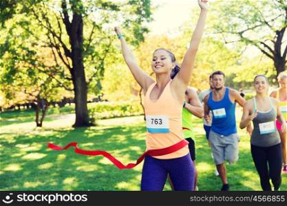 fitness, sport, victory, success and healthy lifestyle concept - happy woman winning race and coming first to finish red ribbon over group of sportsmen running marathon with badge numbers outdoors