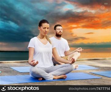 fitness, sport, yoga and healthy lifestyle concept - couple meditating in lotus pose on sea pier over sunset background. couple meditating in yoga lotus pose outdoors