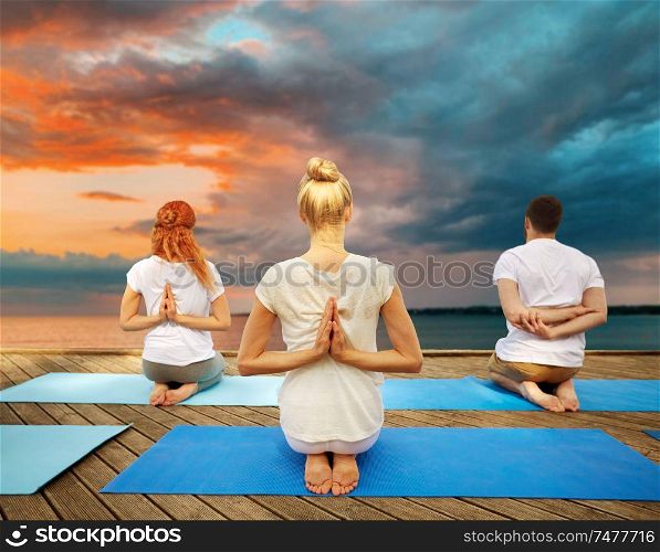 fitness, sport, yoga and healthy lifestyle concept - group of people exercising in reverse prayer pose on sea pier over sunset background. group of people making yoga exercises outdoors