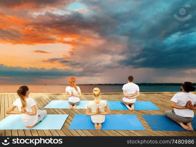 fitness, sport, yoga and healthy lifestyle concept - group of people exercising in reverse prayer pose on sea pier over sunset background. group of people making yoga exercises outdoors