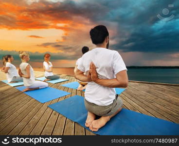 fitness, sport, yoga and healthy lifestyle concept - group of people exercising in reverse prayer pose on sea pier over sunset background. group of people making yoga exercises outdoors