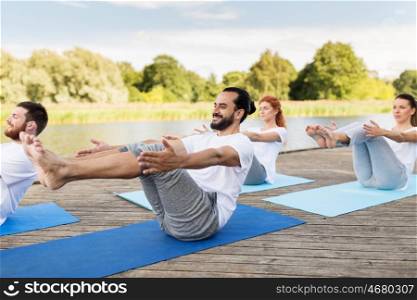fitness, sport, yoga and healthy lifestyle concept - group of people making half-boat pose on mat outdoors on river or lake berth