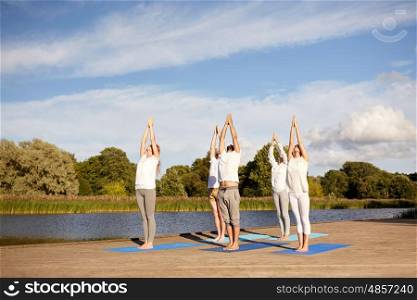 fitness, sport, yoga and healthy lifestyle concept - group of people making upward salute pose on river or lake berth