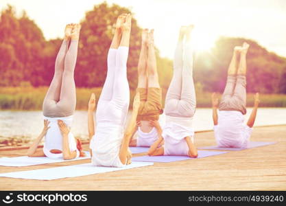 fitness, sport, yoga and healthy lifestyle concept - group of people making shoulderstand pose on river or lake berth. group of people making yoga exercises outdoors