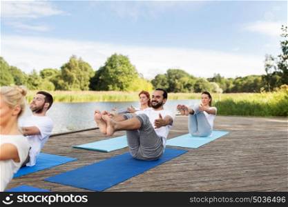 fitness, sport, yoga and healthy lifestyle concept - group of people making half-boat pose on mat outdoors on river or lake berth. people making yoga in half-boat pose outdoors