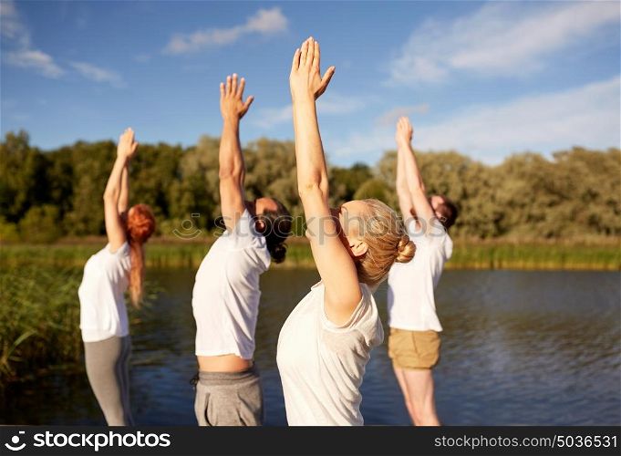 fitness, sport, yoga and healthy lifestyle concept - group of people making upward salute pose on river or lake berth. group of people making yoga exercises outdoors