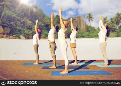 fitness, sport, yoga and healthy lifestyle concept - group of people making upward salute pose over tropical beach background. group of people making yoga exercises over beach